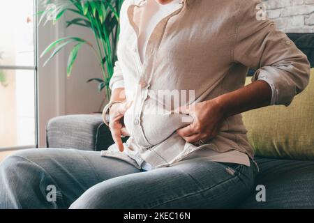 Section médiane des mains de la femme tenant sa graisse du ventre assis sur le canapé à la maison. Une femme caucasienne attrape trop de graisse sur son abdomen. Jeune femme se pinçant sur sa taille grasse obèse Banque D'Images