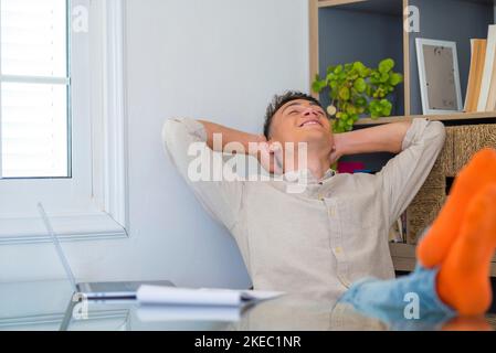 Un adolescent satisfait est heureux de terminer son travail avec un ordinateur portable à la maison, lève les mains et met les pieds sur la table, se détend après une dure journée de travail en prévision d'un congé de fin de semaine, d'une journée de travail détendue, sans stress Banque D'Images