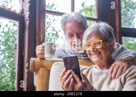 Couple de deux personnes âgées et matures à la maison utilisant le téléphone ensemble dans le canapé. Smartphone pour les personnes âgées, amusant et agréable à regarder. Concept loisir et temps libre dans le salon Banque D'Images
