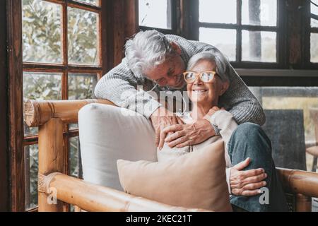 Un vieux couple caucasien passe du temps libre ensemble à la maison. Un mari aimant embrassant la femme de derrière assis sur un fauteuil à la maison. Romantique homme âgé embrassant son partenaire de vie se reposant sur une chaise Banque D'Images