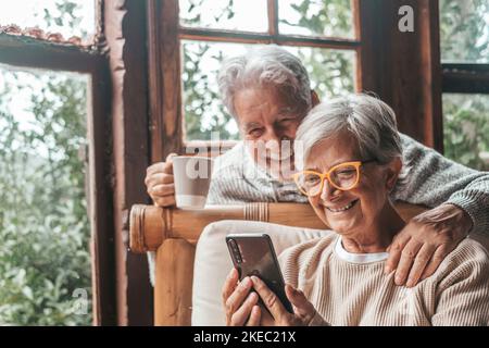 Couple de deux personnes âgées et matures à la maison utilisant le téléphone ensemble dans le canapé. Smartphone pour les personnes âgées, amusant et agréable à regarder. Concept loisir et temps libre dans le salon Banque D'Images