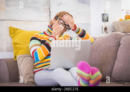 une jeune femme malade et malade se sent mal avec la grippe et un certain virus sur le canapé à la maison.Femme souffrant de fièvre et de maux de tête à l'aide d'un ordinateur portable ou d'un appareil technologique. Banque D'Images