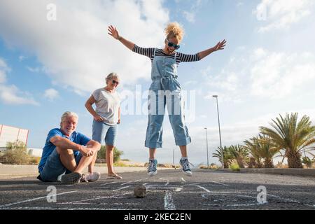 groupe de trois personnes caucasiennes s'amusant et jouant à le hopscotch ensemble - belle femme sautant au milieu de l'asphalte et de deux aînés la regardant Banque D'Images