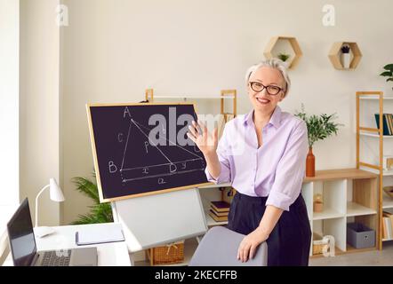 Positive adulte gris cheveux femme enseignant se tient à côté de la table avec ordinateur portable et tableau noir Banque D'Images