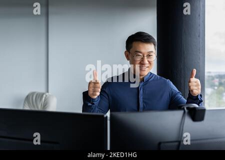 Un jeune homme d'affaires asiatique est assis dans un bureau moderne à un ordinateur. Organise une réunion en ligne par appel vidéo, communique avec les partenaires, pointe les super doigts, sourit. Banque D'Images