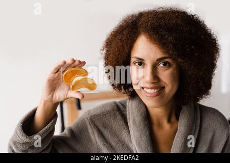 Femme africaine tenant des taches d'or pour lisser les rides, enlever l'enflure ou les sacs sous les yeux. Une fille en peignoir avec des timbres dorés pour les yeux en hydrogel Banque D'Images