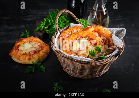 Mini pizza pour enfants, petites tartes ouvertes avec jambon, sauce tomate, mozzarella et fromage dans un panier sur fond sombre. Banque D'Images