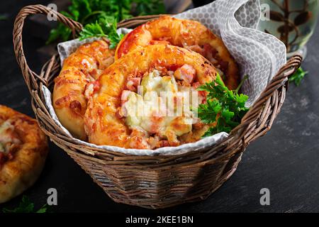 Mini pizza pour enfants, petites tartes ouvertes avec jambon, sauce tomate, mozzarella et fromage dans un panier sur fond sombre. Banque D'Images