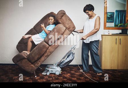 Les grands-parents ont obtenu une super force. Une grand-mère passant l'aspirateur sous un canapé sur lequel son petit-fils est couché à la maison. Banque D'Images