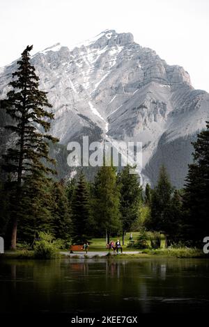 Un couple âgé parle avec un enfant sur des bancs dans le parc au bord de la rivière Banff avec la montagne en arrière-plan Banque D'Images