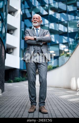 Homme d'affaires souriant en costume qui donne sur l'extérieur tout en se tenant debout avec les bras croisés sur la passerelle piétonne contre le bâtiment du siège social Banque D'Images