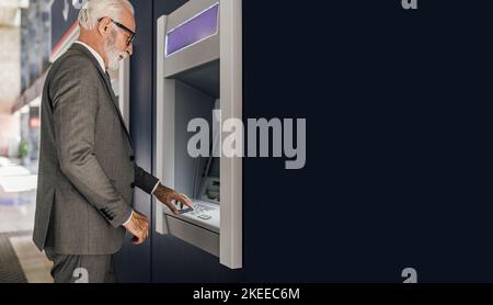 Vue latérale de l'homme d'affaires senior à l'aide d'un distributeur automatique de billets. Le professionnel des personnes âgées portant des lunettes de vue saisit le code PIN pour le retrait d'argent. Il est en costume wh Banque D'Images