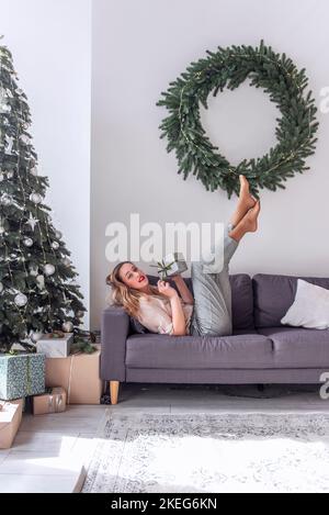 Femme millénaire avec rouge à lèvres en forme de punaise, tenant un cadeau dans les mains, les jambes soulevées sur un canapé gris près de l'arbre de Noël, couronne. Fou Nouveau Banque D'Images