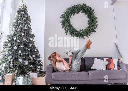 Femme millénaire avec rouge à lèvres en forme de punaise, tenant un cadeau dans les mains, les jambes soulevées sur un canapé gris près de l'arbre de Noël, couronne. Fou Nouveau Banque D'Images