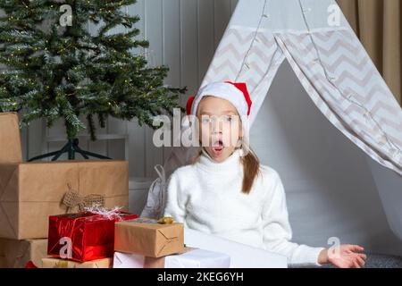 Une fille émotive dans un chapeau de père Noël est assise sur le sol à côté d'une pile de ses cadeaux dans la pépinière. L'enfant est heureux avec des cadeaux de Santa. Le chi Banque D'Images