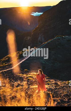 Description: Belle femme en robe rouge souriant dans l'atmosphère de soirée ensoleillée sur Pico do Ariero. Verade do Pico Ruivo, île de Madère, Portugal, E Banque D'Images