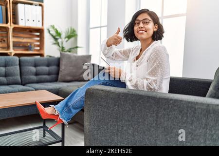 Jeune femme thérapeute hispanique travaillant au bureau de consultation souriant heureux et positif, pouce vers le haut faisant excellent et signe d'approbation Banque D'Images