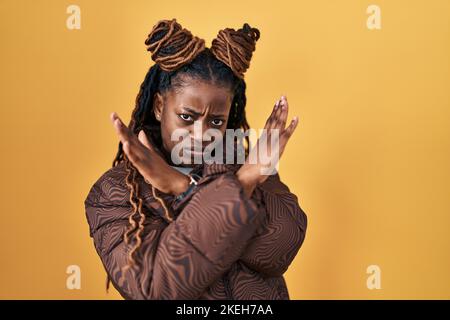 Femme africaine aux cheveux tressés debout sur fond jaune rejet expression croisant les bras faisant signe négatif, visage en colère Banque D'Images