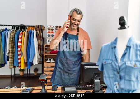 Homme d'âge moyen à cheveux gris assistant de magasin utilisant l'ordinateur parlant sur le smartphone à la boutique de vêtements Banque D'Images