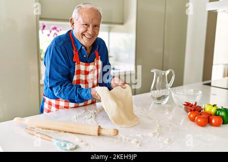 Homme âgé souriant confiant tenant la pâte avec les mains dans la cuisine Banque D'Images