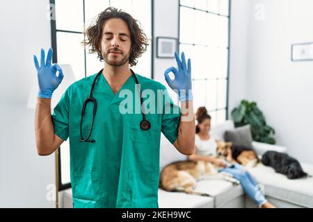 Jeune vétérinaire homme vérifiant les chiens à la maison se détendre et sourire avec les yeux fermés faisant le geste de méditation avec les doigts. Concept de yoga. Banque D'Images