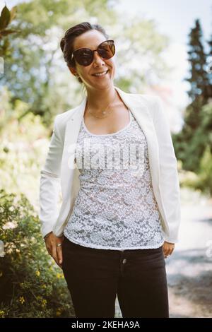Belle jeune bonne femme d'affaires asiatique multiraciale dans blazer blanc, chemise en dentelle, jeans et lunettes de soleil dans le parc d'été, souriant Banque D'Images