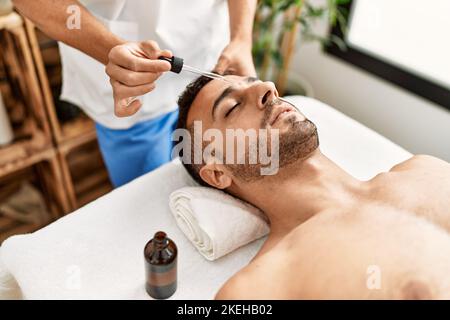 Deux hommes hispaniques thérapeute et patient ayant un traitement du visage en utilisant le sérum au centre de beauté Banque D'Images