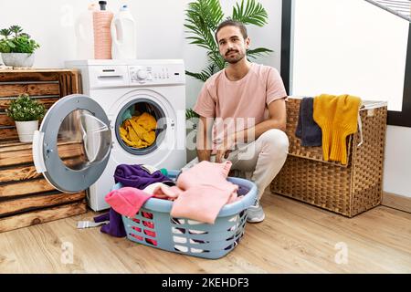 Jeune hispanique mettant le linge sale dans la machine à laver avec une expression sérieuse sur le visage. Simple et naturel regarder la caméra. Banque D'Images