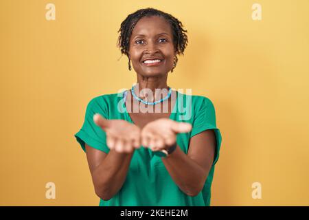 Femme africaine avec des dreadlocks debout sur fond jaune souriant avec les mains paumes ensemble recevoir ou donner le geste. Tenir et la protection Banque D'Images