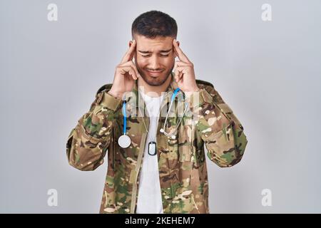 Jeune médecin hispanique portant l'uniforme militaire de camouflage avec main sur la tête, maux de tête parce que le stress. Souffrant de migraine. Banque D'Images