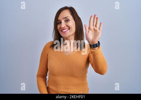 Femme brune d'âge moyen debout portant un chandail orange en train de dire bonjour heureux et souriant, geste de bienvenue amical Banque D'Images