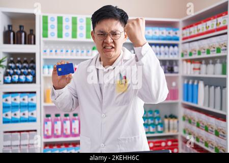 Un jeune homme asiatique travaillant dans une pharmacie en pharmacie qui tient une carte de crédit agacé et frustré criant avec colère, hurlant fou de colère et de main levée Banque D'Images