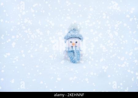 Adorable bonhomme de neige en peluche dans un chapeau bleu et une écharpe sur fond enneigé. Carte de vœux de Noël et du nouvel an. Concept d'hiver. Copier l'espace. Banque D'Images