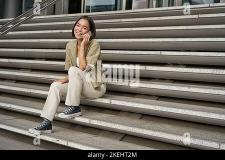 Portrait d'une jeune fille étudiante asiatique dans les escaliers parle sur un téléphone portable, sourit à l'appareil photo, se trouve à l'extérieur du bâtiment Banque D'Images