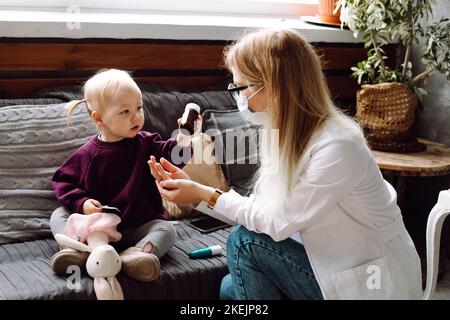 Portrait d'une merveilleuse petite fille assise sur un canapé, donnant une bouteille brune au médecin des enfants jouant avec le patient à la maison. Banque D'Images