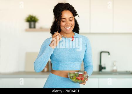 Jeune femme sportive afro-américaine mangeant de la salade de légumes frais dans la cuisine, appréciant la nourriture pleine de vitamines, l'espace de copie Banque D'Images