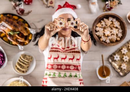 Une cuisinière gaie dans un tablier de Noël se trouve sur le sol et couvre ses yeux avec des gâteaux, entouré de plats de vacances traditionnels et des gâteaux. Banque D'Images