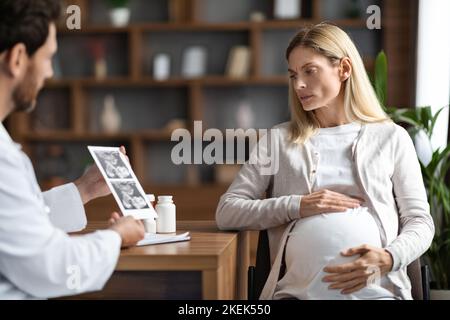Gynécologue masculin médecin montrant l'image de l'échographie de bébé à la femme enceinte inquiète Banque D'Images
