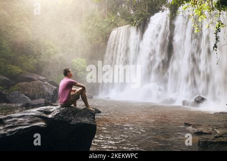 Homme assis sur le rocher devant la haute chute d'eau dans les montagnes dans le paysage tropical à Camobodia. Banque D'Images