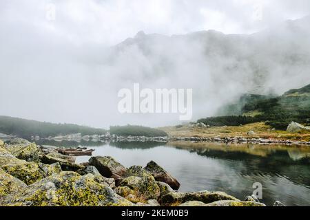 Lac de montagne clair avec réflexion encerclée par des roches mossy et des pins nains sur la rive du lac avec haute montagne couverte par brouillard épais dans les Tatras polonais Banque D'Images