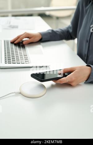 Gros plan d'une femme d'affaires qui connecte son smartphone à un chargeur sans fil pour le recharger tout en étant assis à une table au bureau Banque D'Images