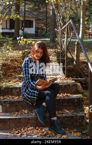 Une jolie jeune dame lit un livre sur les escaliers au coucher du soleil dans la forêt d'automne. Paysage d'automne coloré. Saison d'automne. Serbie, Europe. Banque D'Images