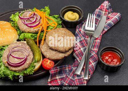 Sauce tomate et moutarde dans des bols. Couteau et fourchette sur serviette. Petits pains ronds, hamburgers frits, cornichons, oignons et laitue sur l'assiette. Arrière-plan noir. Banque D'Images