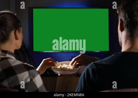 Couple de détente sur le canapé à la maison manger du pop-corn et regarder la télévision. Écran vert vierge Banque D'Images
