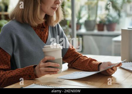 Une jeune femme d'affaires boit du café à partir d'une tasse jetable tout en étant assise à table et en lisant le document Banque D'Images