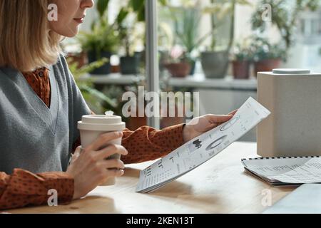 Gros plan de la jeune femme qui boit du café et lit un document avec des graphiques tout en étant assise à table au bureau Banque D'Images