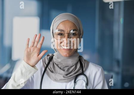 Portrait d'une jeune belle femme, une musulmane, femme arabe médecin. Elle se tient vêtue d'un hijab, de lunettes et d'un stéthoscope, regarde et fait des vagues à la caméra, sourit, salue. Banque D'Images