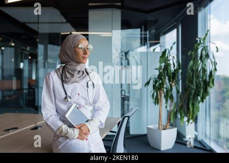 Une jeune femme musulmane heureuse et réfléchie, médecin arabe à l'hôpital, est assise à son bureau, tenant une tablette entre ses mains, regardant la fenêtre, souriant, aimant son travail. Banque D'Images