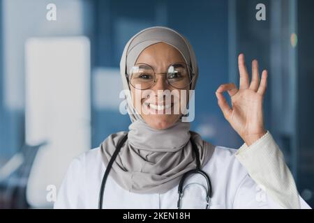 Portrait d'une jeune, belle femme, arabe, médecin musulman. Elle se tient vêtue d'un hijab, de lunettes et d'un stéthoscope, regarde la caméra, sourit, montre avec sa main ok. Banque D'Images