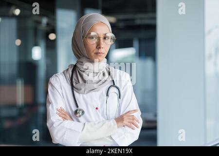 Portrait d'une jeune femme musulmane médecin. Elle est debout dans le hall de l'hôpital vêtu d'un hijab, portant des lunettes et un stéthoscope, regardant sérieusement dans la caméra, ses bras croisés. Banque D'Images